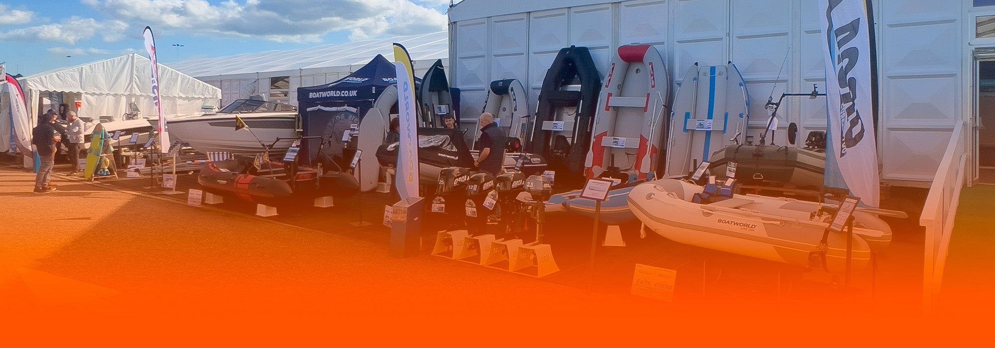 Outdoor marine exhibition stand featuring Boatworld products including inflatable boats, outboard motors and watersports gear, displayed in front of branded tents and banners under a partly cloudy sky.