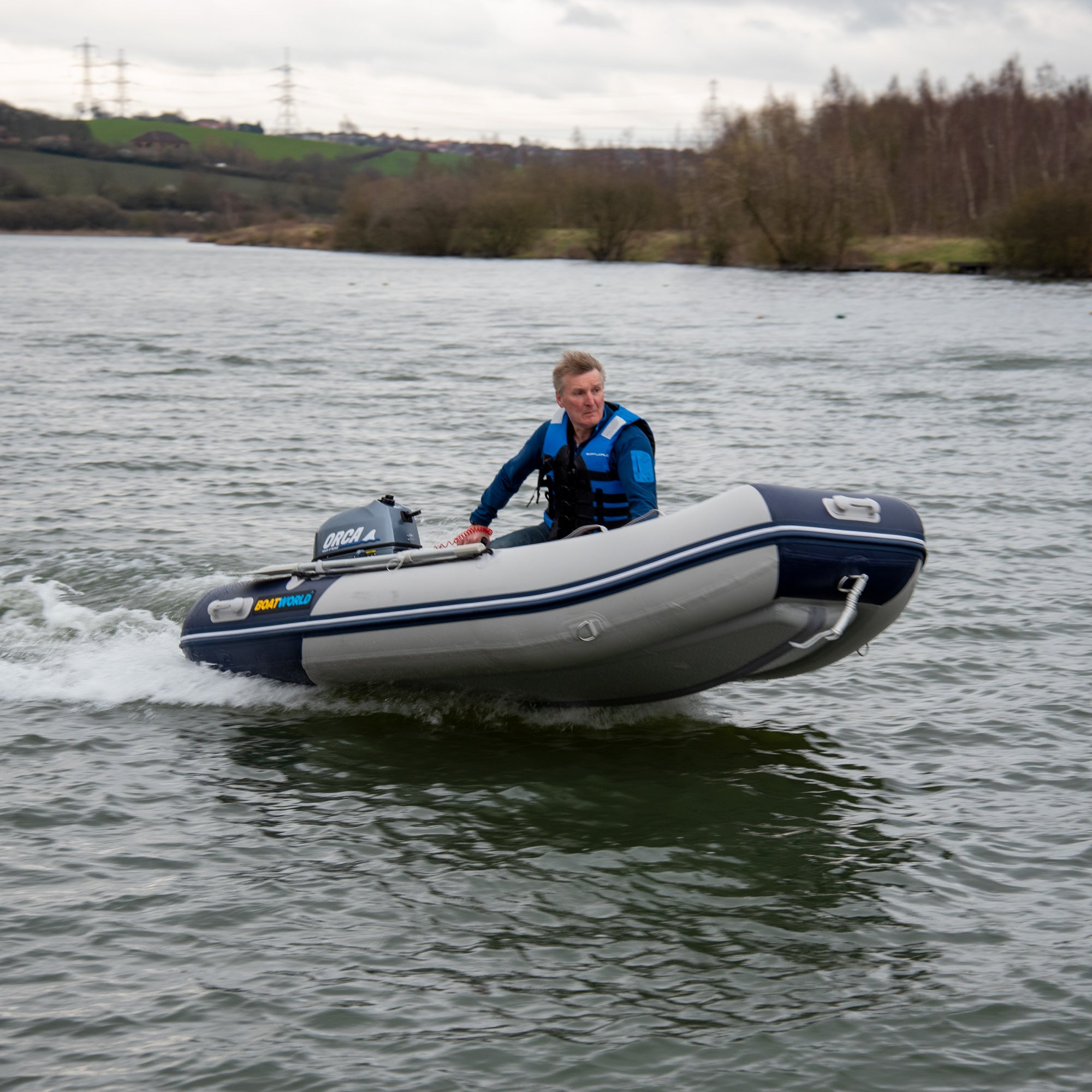 Man in a blue and black life vest piloting a Boatworld Air V-Type 300 small inflatable boat in grey and navy. The boat features an air-filled V-type hull for improved performance and is powered by an Orca outboard motor. It glides across calm water with wooded banks and green hills in the background.