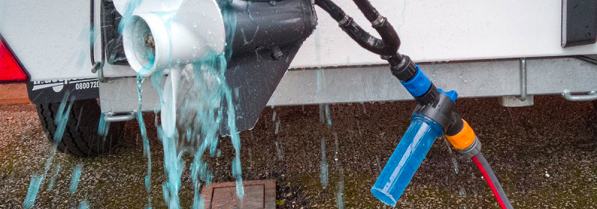 Close-up of an outboard motor being flushed with blue-tinted water using a Blu Thru flushing system, connected to a hose and attached to the motor’s lower unit. Water flows out around the propeller, with a trailer and pavement visible in the background.