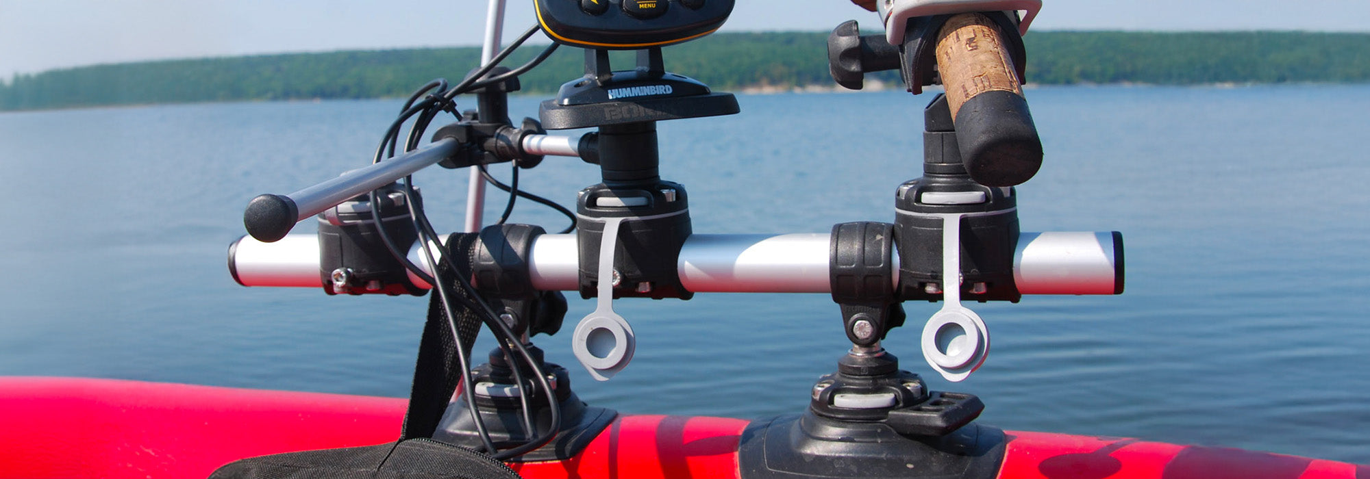Close-up of a fishing setup mounted on a red small inflatable boat, featuring a Borkia aluminium accessory bar with adjustable clamps holding a rod holder and a Humminbird fishfinder. Calm water and a tree-lined shoreline are visible in the background.