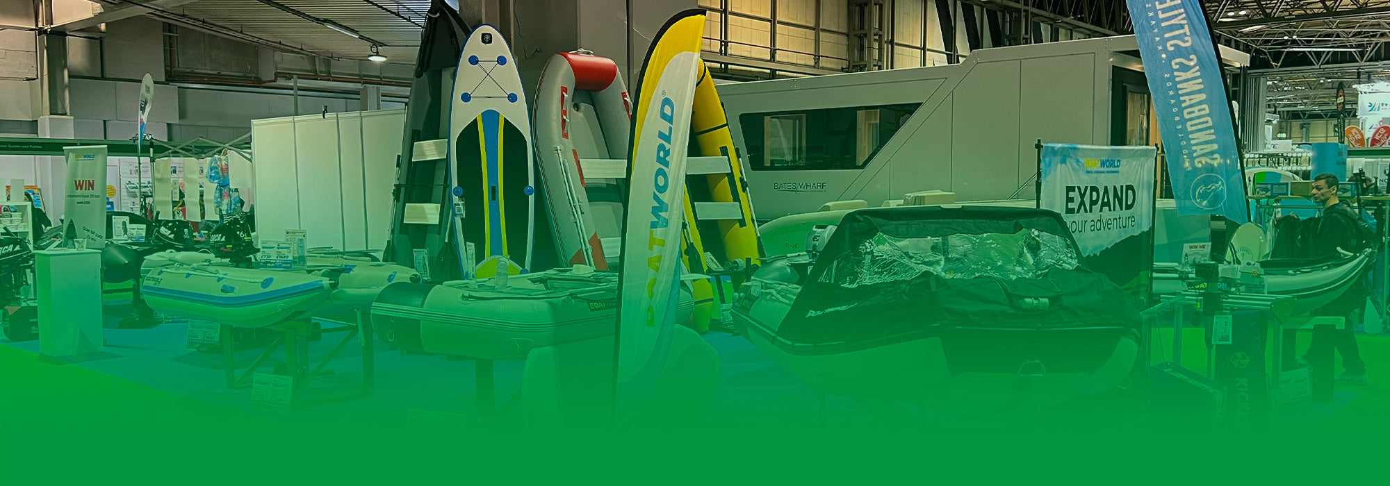 The image shows the Boatworld stand at the Caravan, Camping & Motorhome Show, featuring inflatable boats, paddleboards, and outboard motors. Boatworld-branded banners and a competition sign are visible, along with a blue carpeted floor and bright exhibition lighting.