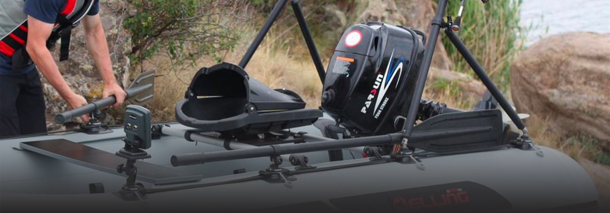Close-up of a small inflatable boat fully outfitted with Borkia accessories, including the a frame, rod holders, fishfinder mount, baiting device, and oar supports. A Parsun outboard motor is mounted at the stern, and a person in a life jacket adjusts an oar beside a rocky, grassy shoreline.