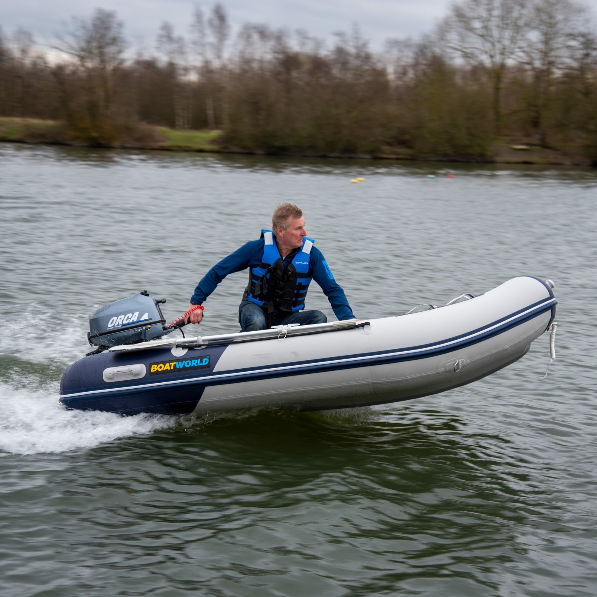 Man in a blue jacket and black life vest steering a Boatworld Air V-Type 300 small inflatable boat across a lake. The grey and navy boat features an air-filled V-type hull and is powered by an Orca outboard motor, with water spray trailing behind and bare trees lining the shore.