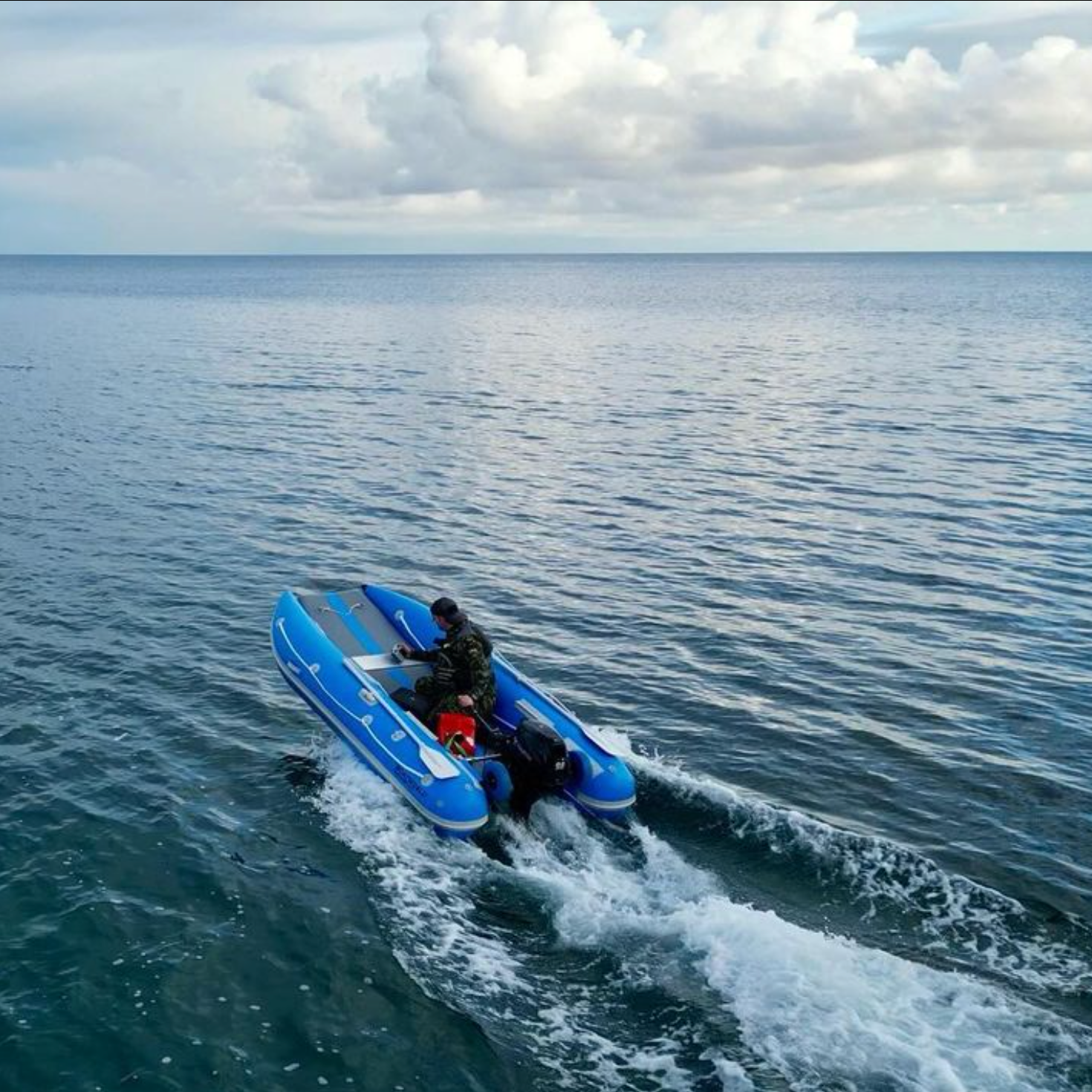 The image features the Boatworld KAT Sport 330, a catamaran-style inflatable boat, moving across open water. A person in camouflage clothing and a cap is steering the boat, which is powered by an outboard motor. The blue inflatable hull cuts through the water, leaving a trail of white foam. The surrounding water is calm with gentle ripples, and the sky is partly cloudy, creating a dynamic and adventurous scene.