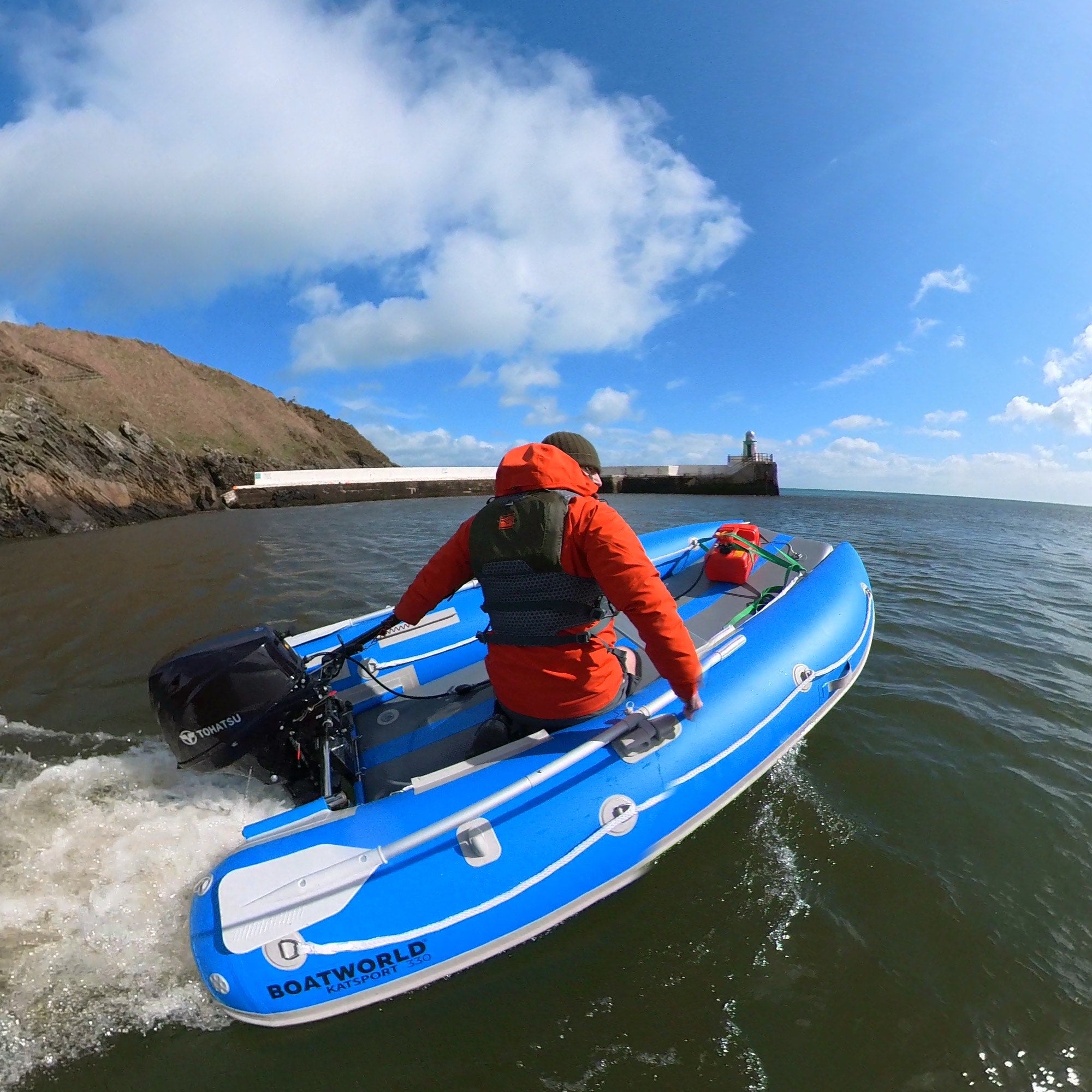 Matt from Airborne Pirates, wearing a red jacket and life vest, pilots a Boatworld KATSPORT 330 catamaran small inflatable boat powered by a Tohatsu outboard motor. The bright blue twin-tube boat cruises along coastal waters, with a rocky shoreline and lighthouse in the distance under a clear blue sky.