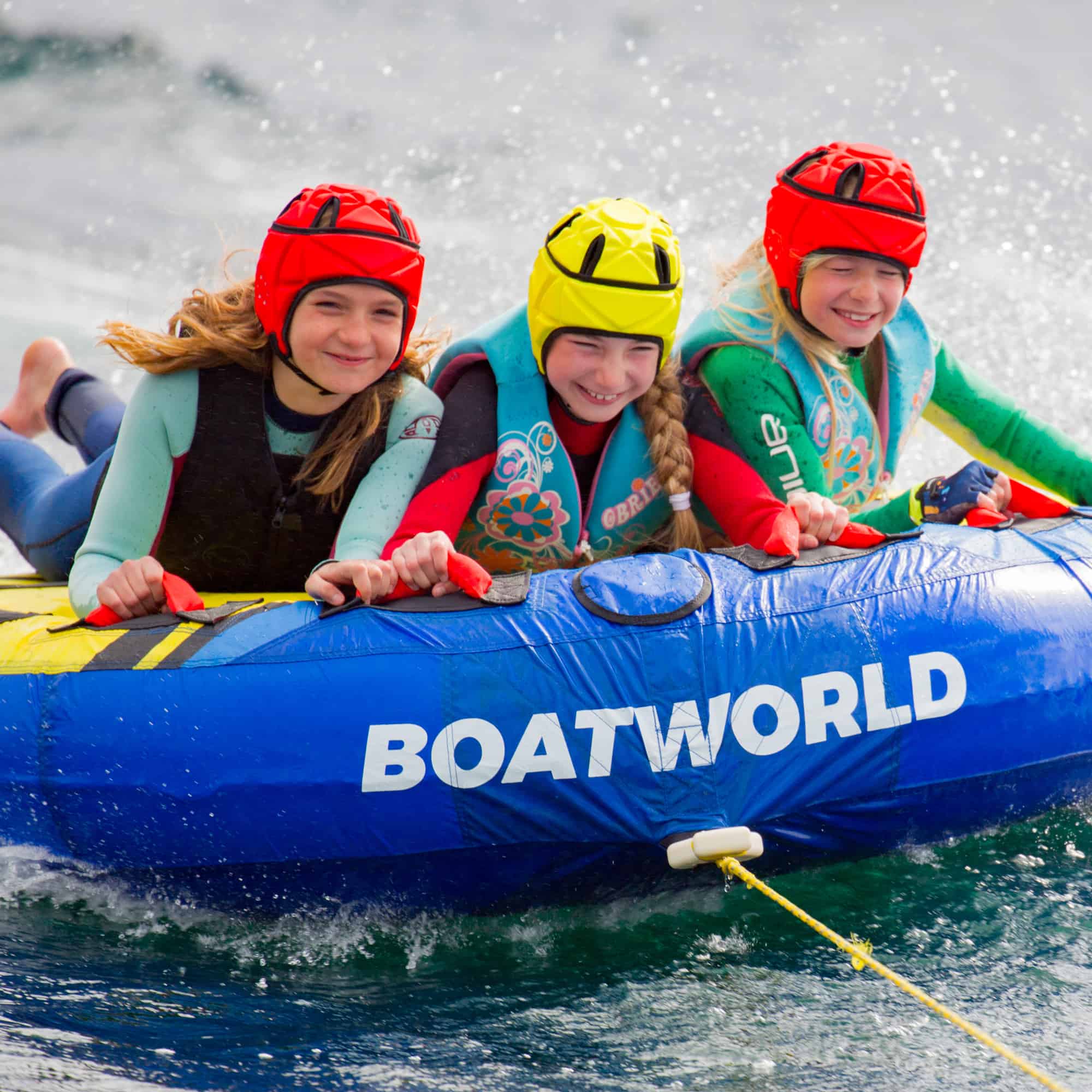 The image features the Boatworld Super Skimmer Inflatable Towable, being ridden by three smiling children wearing helmets and life jackets. The bright blue towable with Boatworld branding is gliding across the water, creating a fun and exciting scene. The riders are gripping the handles tightly as water splashes around them. The vibrant colours of their wetsuits and safety gear contrast with the dynamic movement of the towable, capturing the thrill of the ride.